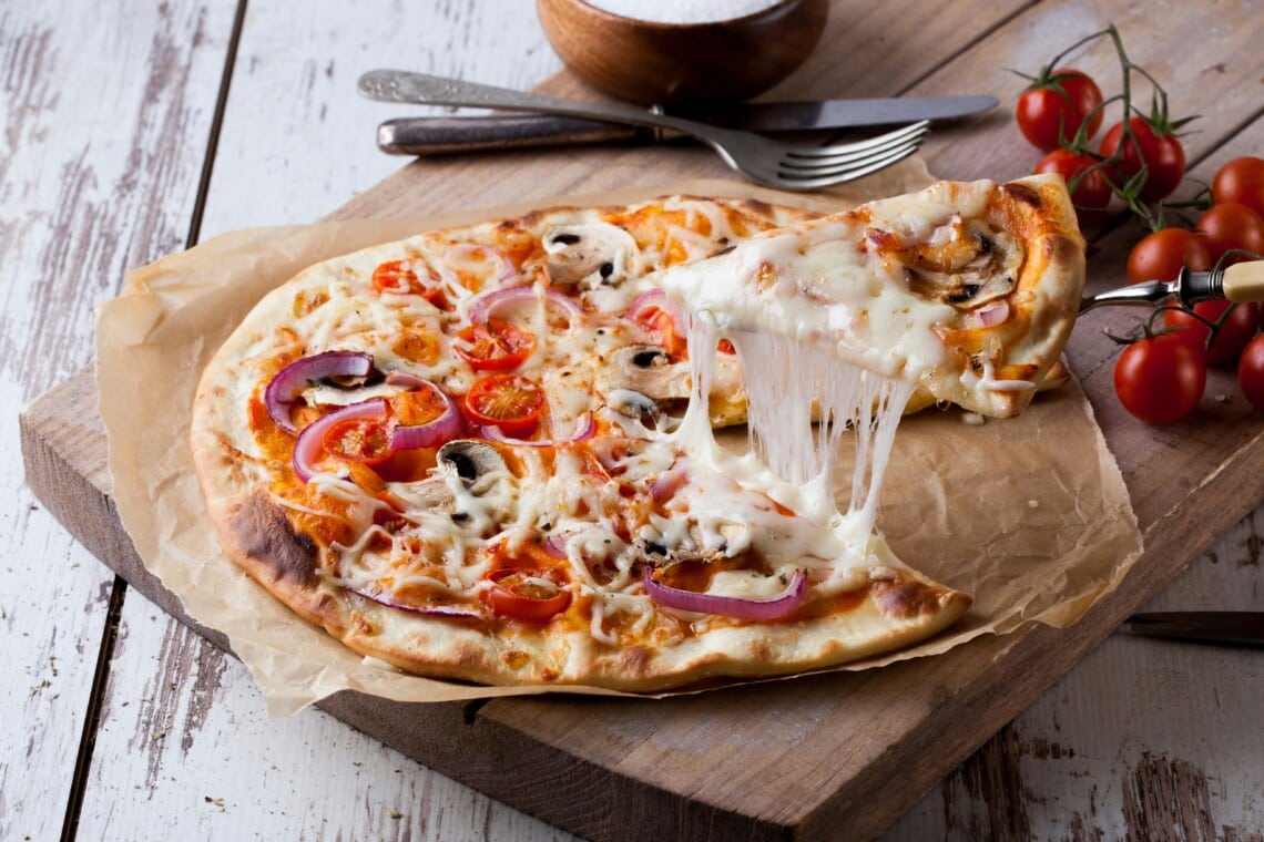 A freshly baked thin-crust pizza topped with cherry tomatoes, red onions, mushrooms, and melted cheese, with one slice being lifted, revealing gooey, stretchy cheese.