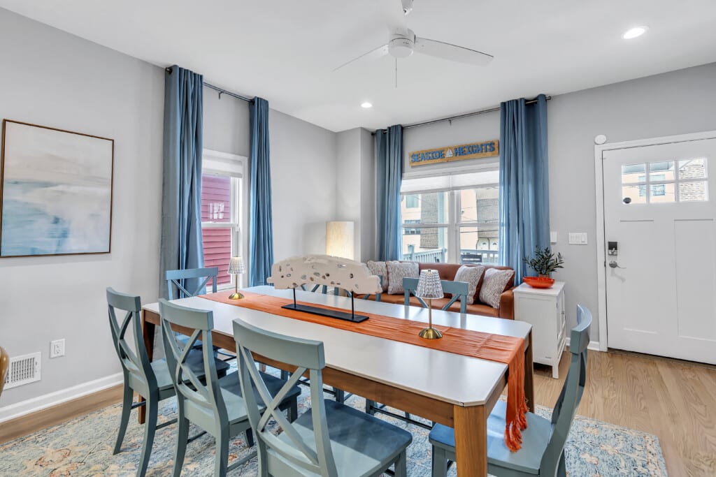 Dining area with brown bed, white windows with blue curtains and a beautiful framed see drawing on the wall.