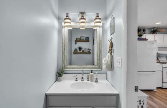 View facing bathroom mirror and sink with hand soap, and kitchen fridge visible next door.