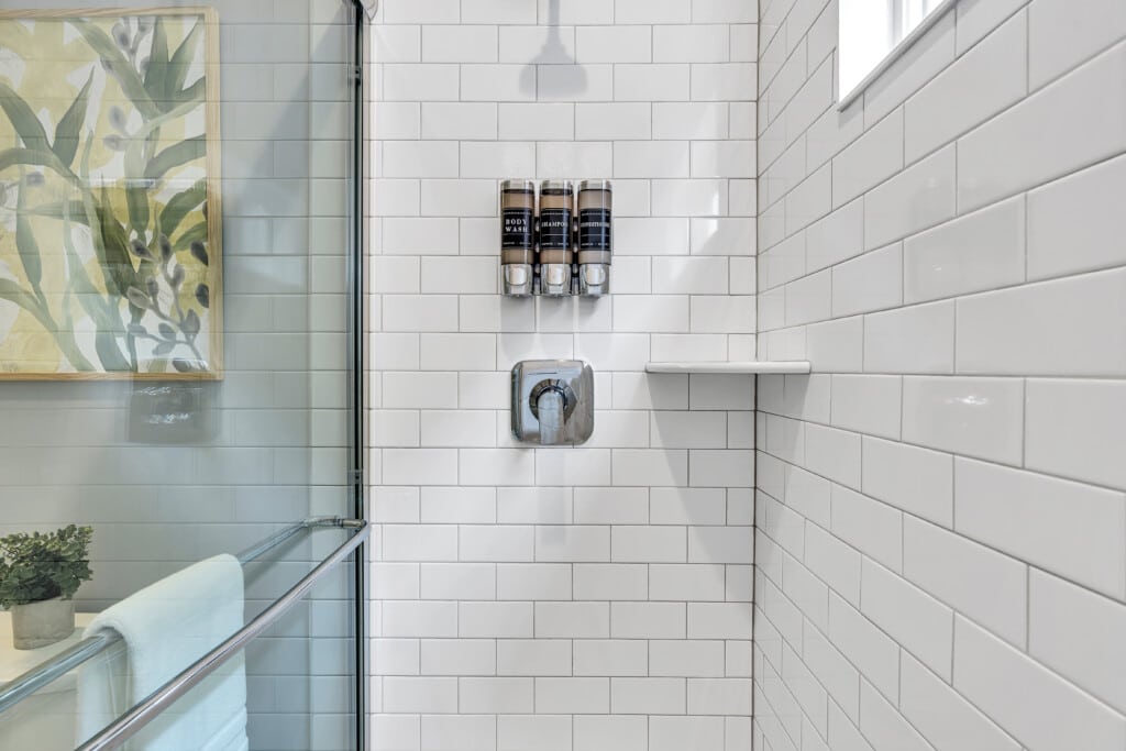 Close-up of white shower tiles with three shampoo bottles hanging inside, whit a beautiful framed picture on the wall.