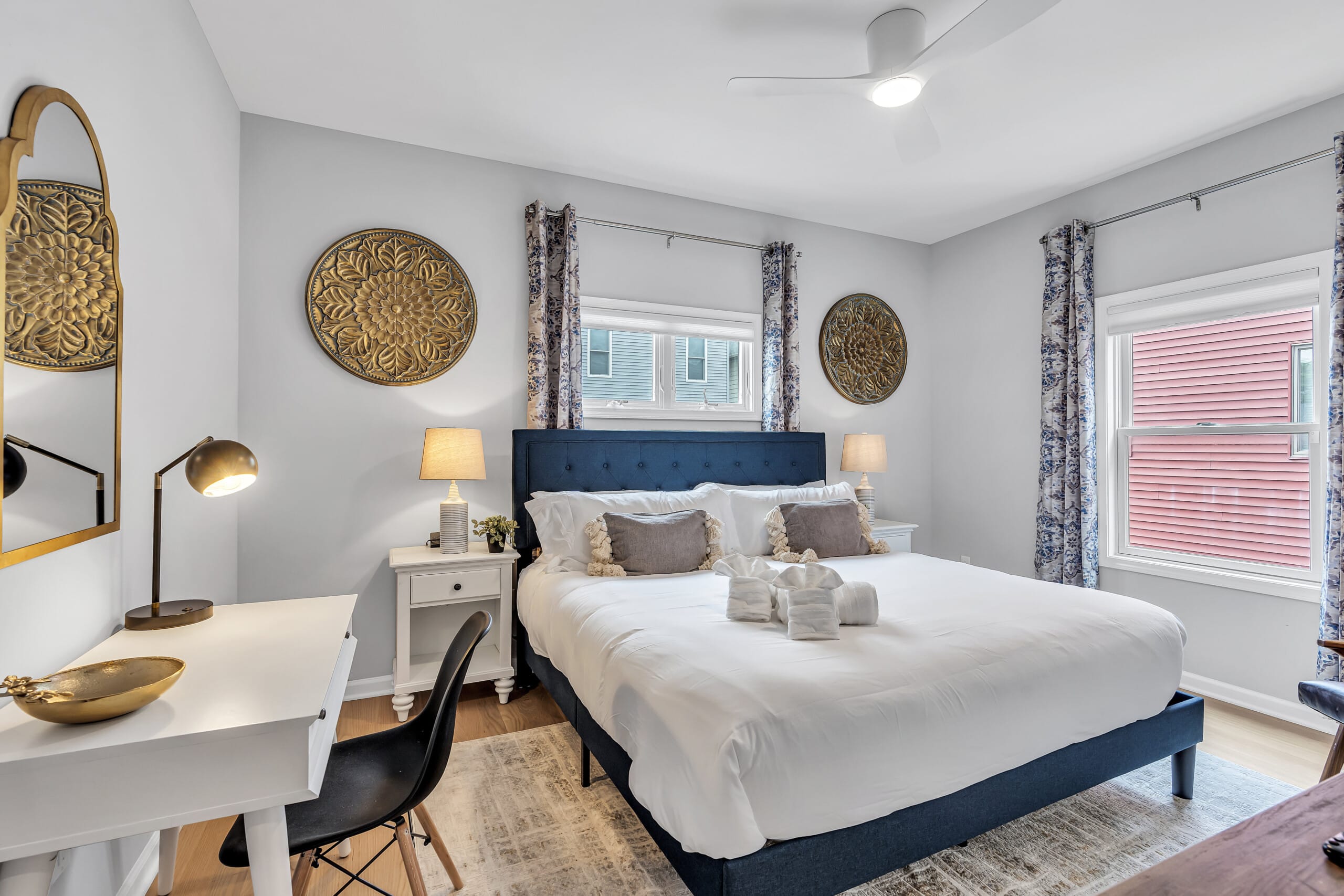 Bedroom with white bedcover, two grey pillows, window with curtains, lamps on each side of the bed, and two framed pictures on the wall.
