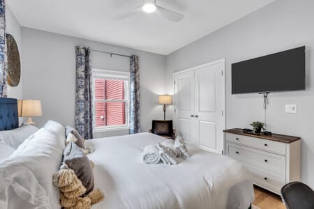 Bedroom with white-covered bed, wall-mounted TV. and white closet door opposite the bed, window with blue curtains.