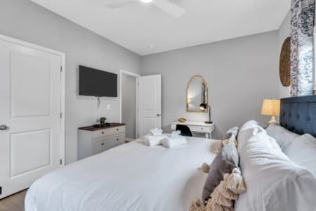 Bedroom with white-covered bed, wall-mounted TV, small table and chair, mirror above table, white door opposite the bed, and lamp on the corner.