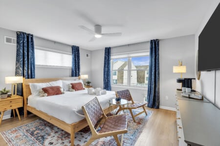Bedroom with white bedcover, two red pillows, window with blue curtains, lamp next to bed, and two chairs and table in front of the bed, tv on the wall.