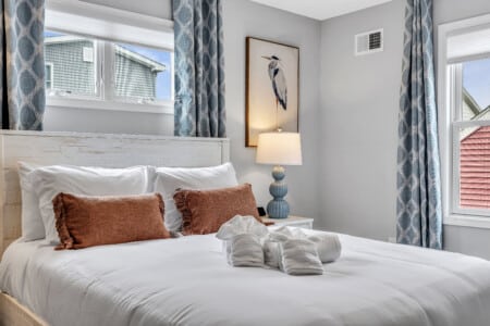 Bedroom with white bedcover, two red pillows and a towel on the bed, window with blue curtains, lamps on each side, and framed picture on the wall.