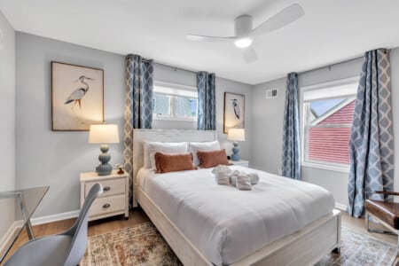 Bedroom with white bedcover, two red pillows, window with blue curtains, lamps on each side, and two framed pictures on the wall.