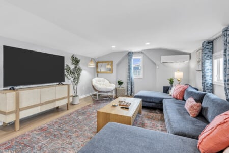 Cozy living room featuring blue sofa, red cushions, white armchair with golden mirror above, table with TV and shells, blue decorative curtains, and green plant in the corner.