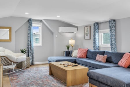 Living room with blue couch, red pillows, comfy white chair, table with shell decor, and windows with blue curtains.