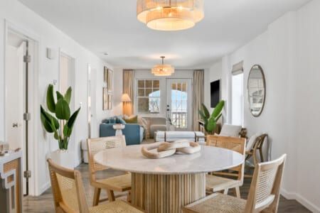 Cozy living room with balcony view, comfy blue couch, two white chairs, circle mirror, open window, and dining table.