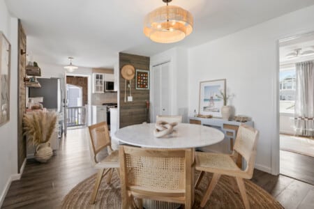 Dining room with table, white kitchen, balcony view, chandelier, and side table holding plant and framed photo.