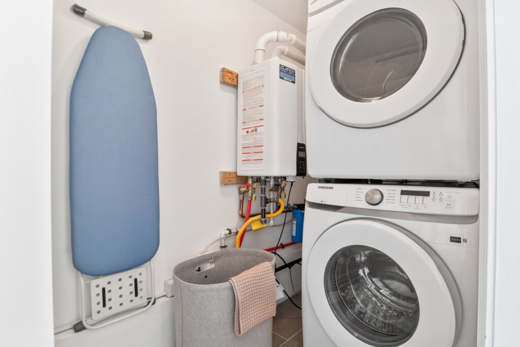 Small laundry room with dryer and washing machine, blue ironing board on wall, and grey laundry box on floor.