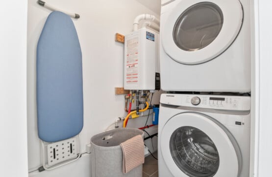 Small laundry room with dryer and washing machine, blue ironing board on wall, and grey laundry box on floor.