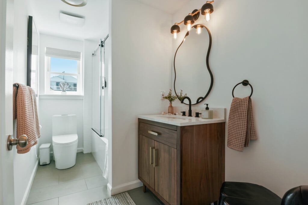 Bathroom with toilet under window, shower, sink with black tap, light brown towel, and mirror with lights above.