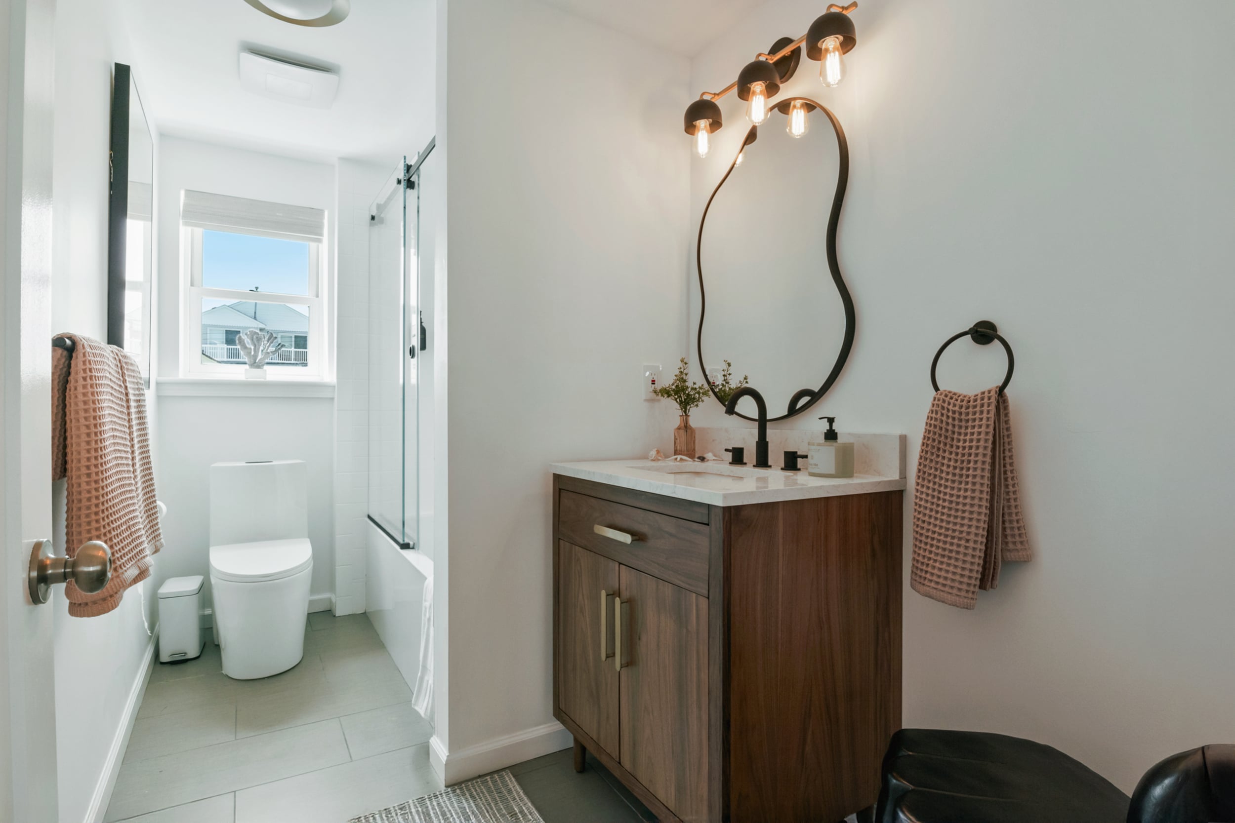 Bathroom with toilet under window, shower, sink with black tap, light brown towel, and mirror with lights above.