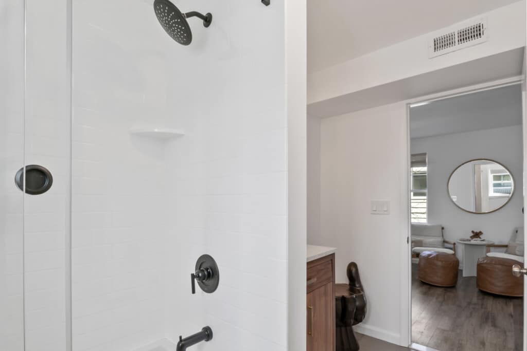 Half view of bathroom with white tiles and black shower, and two white chairs with round mirror above.