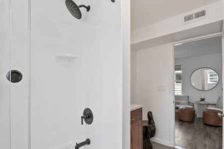 Half view of bathroom with white tiles and black shower, and two white chairs with round mirror above.