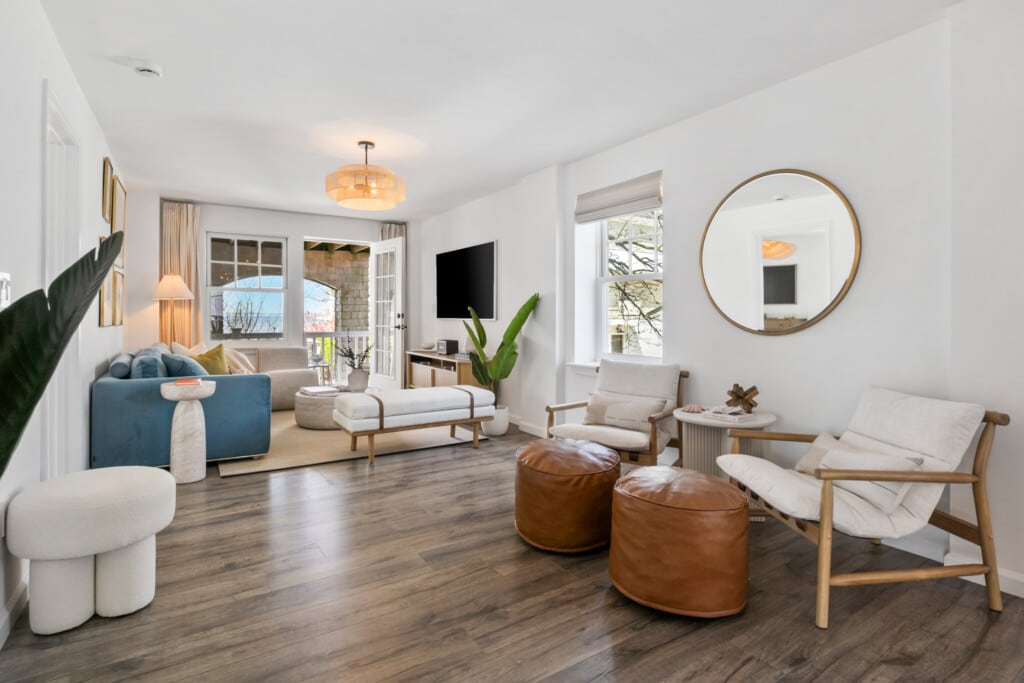 Cozy living room with balcony view, comfy blue couch, two comfy white chairs with a circle mirror above them and white open window.