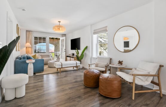 Cozy living room with balcony view, comfy blue couch, two comfy white chairs with a circle mirror above them and white open window.