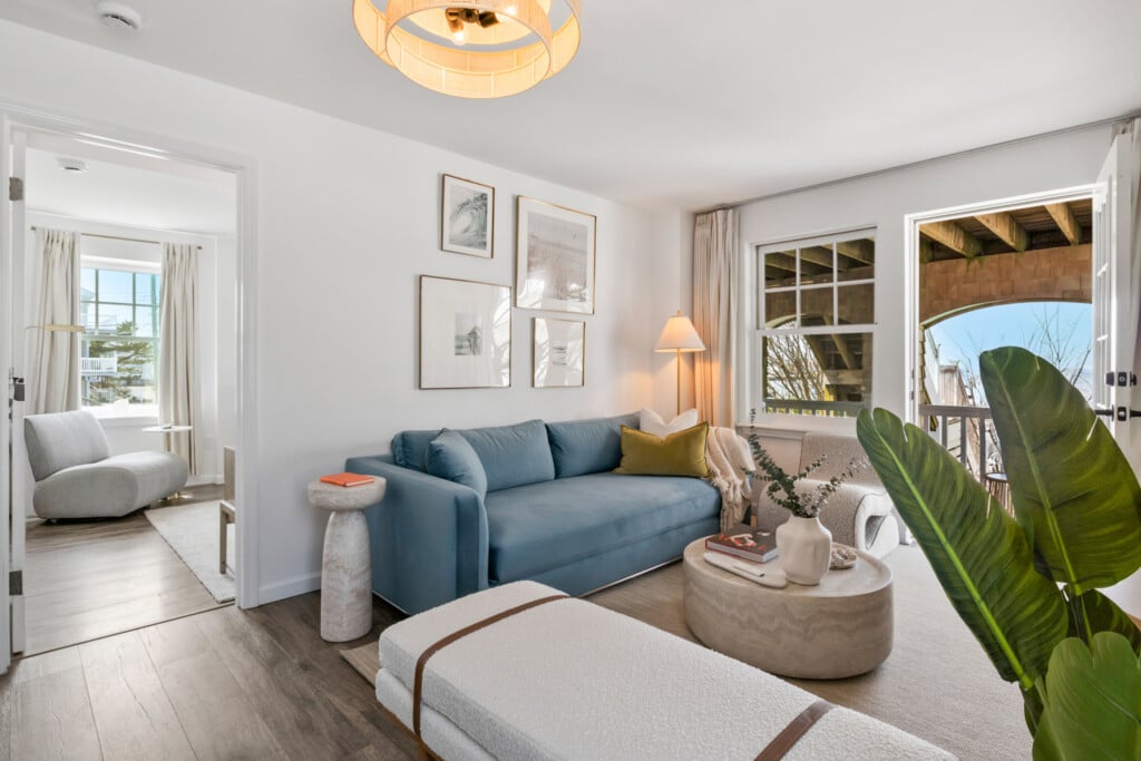 Living room, blue couch, white sofa, beautiful pictures on the wall, lighting lamp in the corner and a table holding a vase and a book, grey sofa in the separate room.