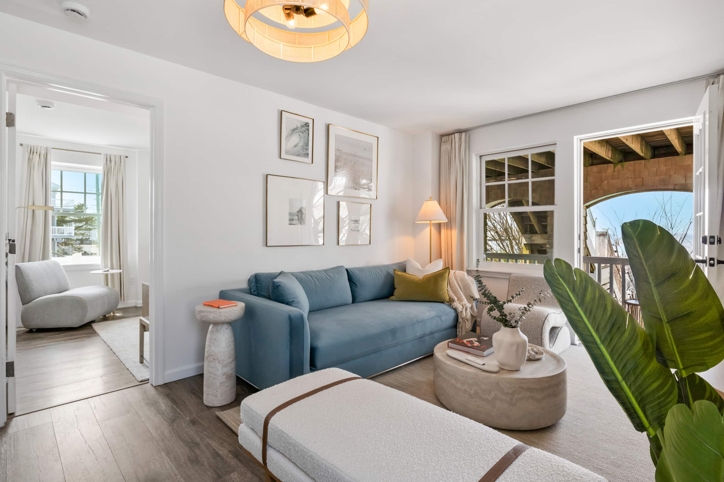Living room, blue couch, white sofa, beautiful pictures on the wall, lighting lamp in the corner and a table holding a vase and a book, grey sofa in the separate room.