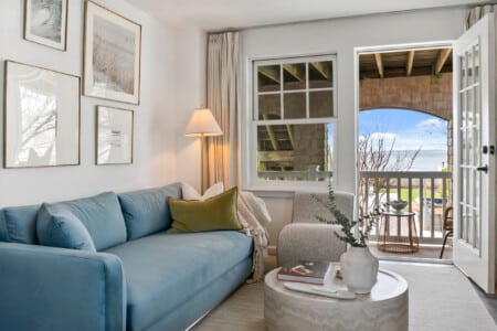 Living room with a balcony view, blue couch, white sofa, beautiful pictures on the wall, lighting lamp in the corner and a table holding a vase and a book.