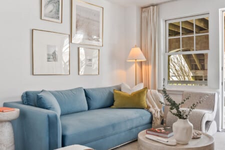 Living room, blue couch, white sofa, beautiful pictures on the wall, lighting lamp in the corner and a table holding a vase and a book.