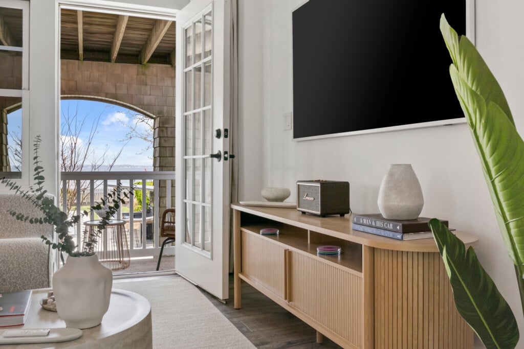Room with TV on wall, table with radio, books, and vases, plus a view of the outside with a table holding green plant.