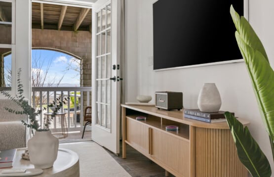 Room with TV on wall, table with radio, books, and vases, plus a view of the outside with a table holding green plant.