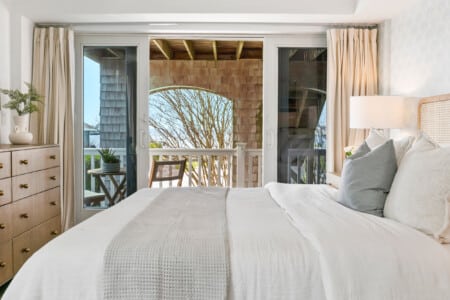 Cozy bedroom with white bedding, sunset view, beige lamp in the corner, and balcony with chairs and a table.