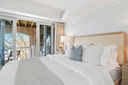 Cozy bedroom with white bedding, bedside table holding a lamp and plant, and views of the balcony and sunset.