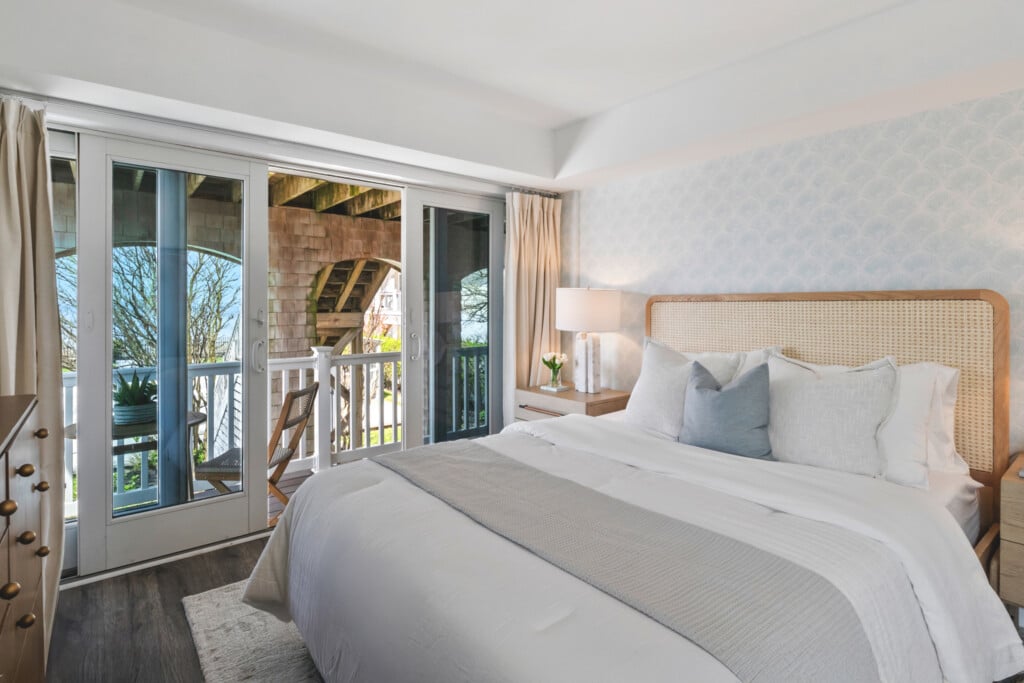 Bedroom with white bed and pillows, table with lamp and plant vase, plus balcony and sunset views outside.