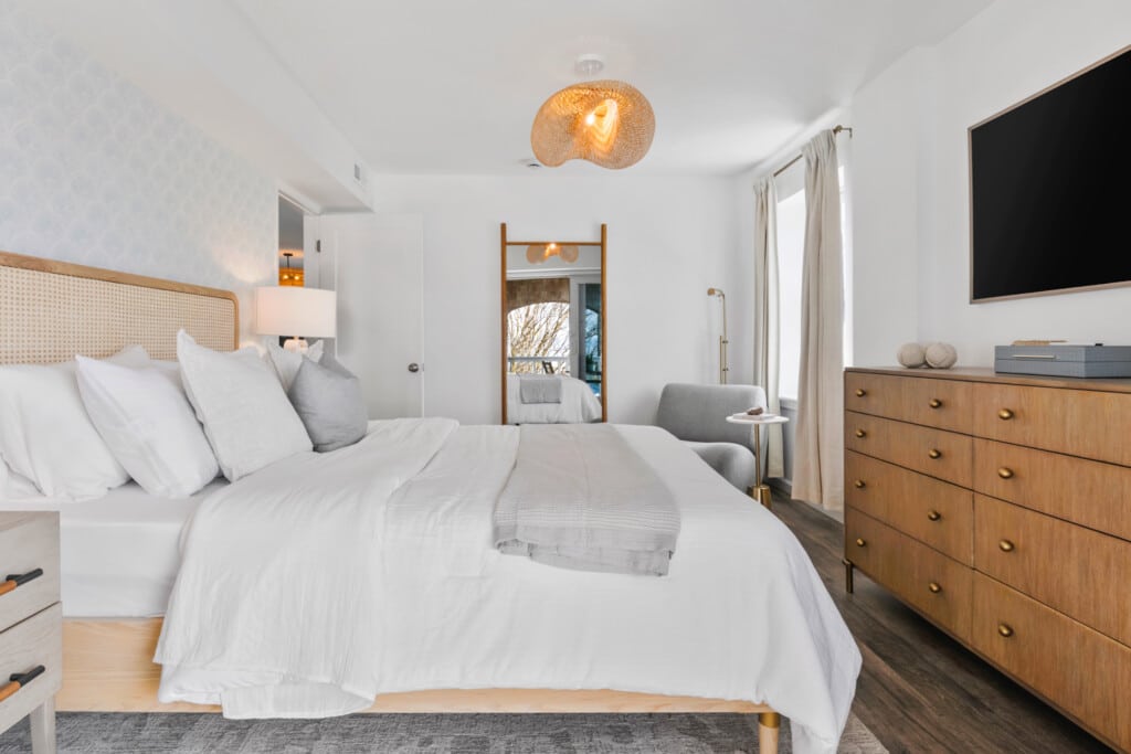 Bedroom with white bed, grey sofa, large wall mirror, and TV mounted opposite the bed.