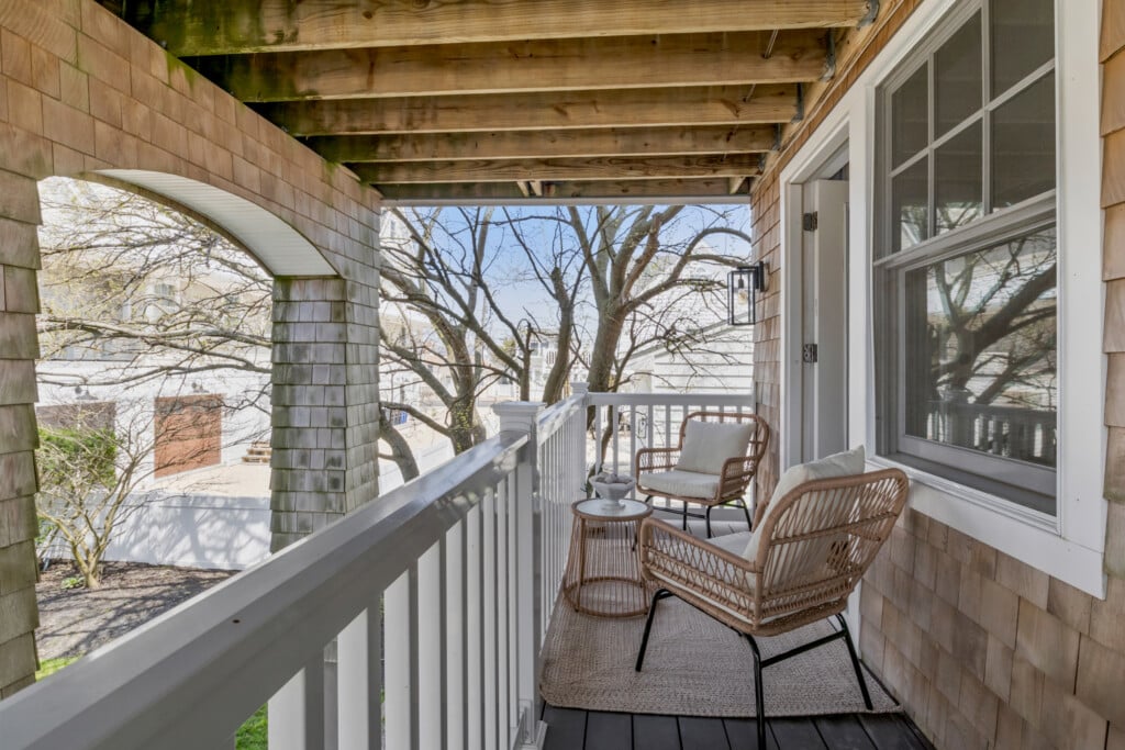 Cozy covered balcony with wicker chairs and a small table, overlooking trees and a quiet residential street.