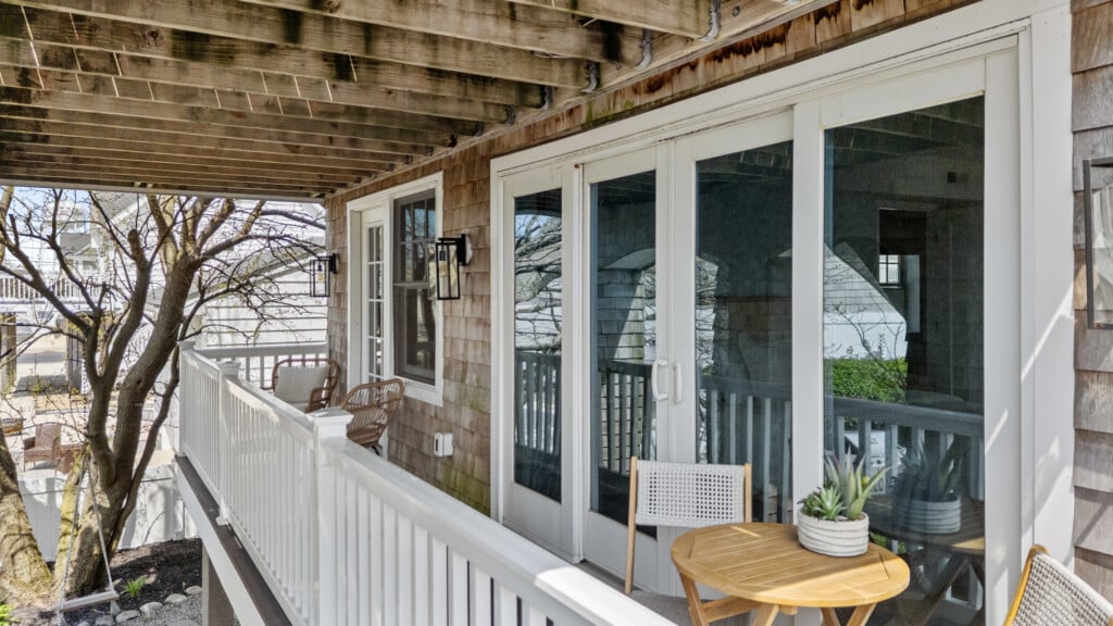 Sunny balcony with white walls, two tables with chairs, white doors and windows, and a plant on one table.