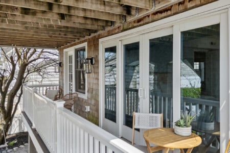 Sunny balcony with white walls, two tables with chairs, white doors and windows, and a plant on one table.
