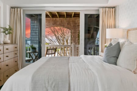Cozy bedroom with white bedding, sunset view, beige lamp in the corner, and balcony with chairs and a table.