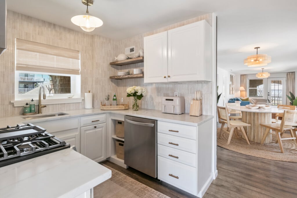 White kitchen with window, grey dishwasher, table with white flowers and water tray, and living room view.