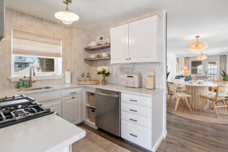 White kitchen with window, grey dishwasher, table with white flowers and water tray, and living room view.