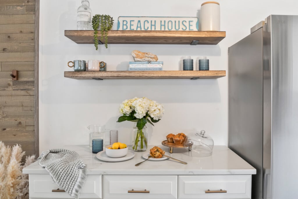 Table with water glass, muffins, lemons, white flowers, grey fridge, and a “Beach House” decoration.