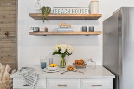 Table with water glass, muffins, lemons, white flowers, grey fridge, and a “Beach House” decoration.