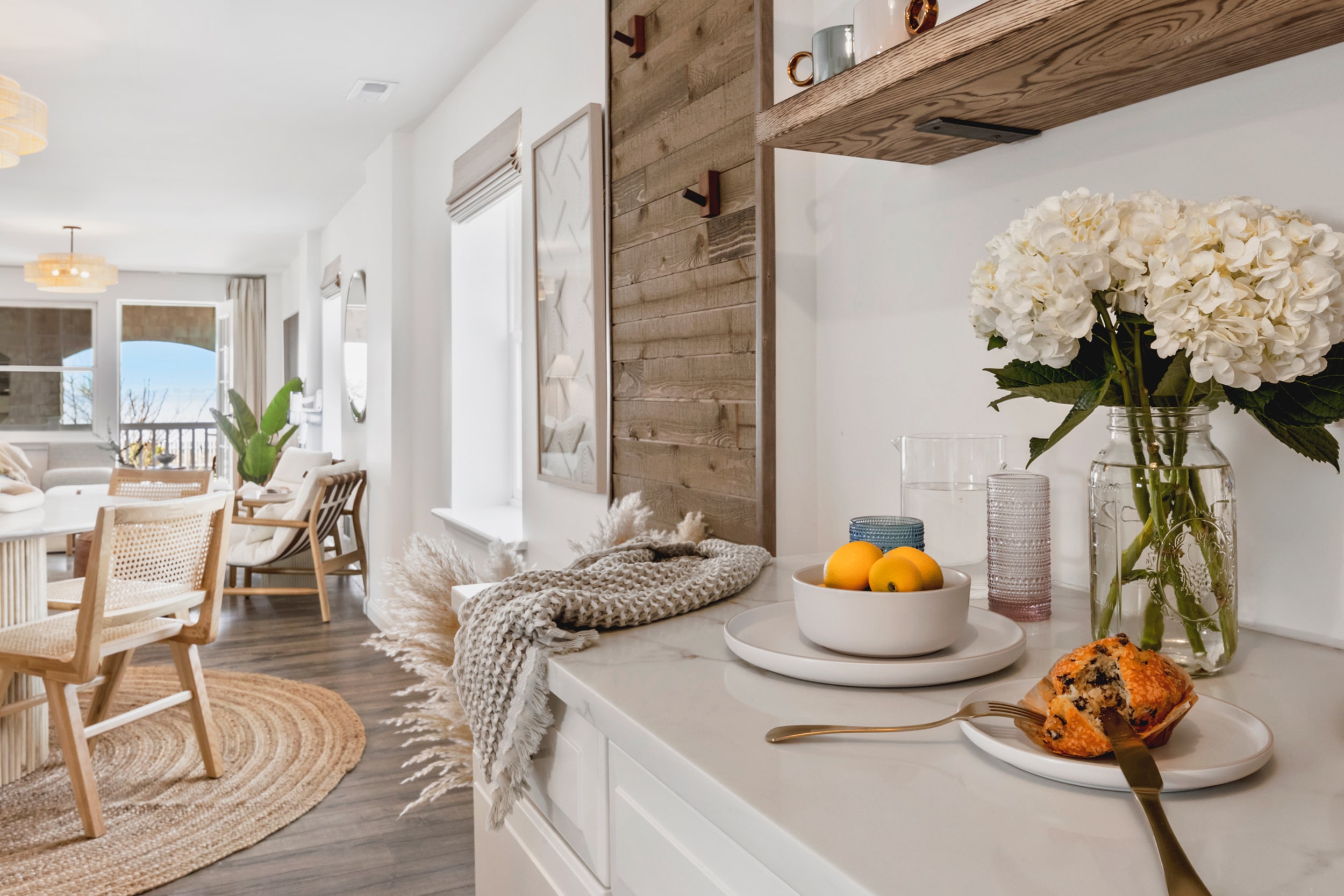 White table with glass of water, muffins, plate of lemons, and vase with white flowers and living room view.