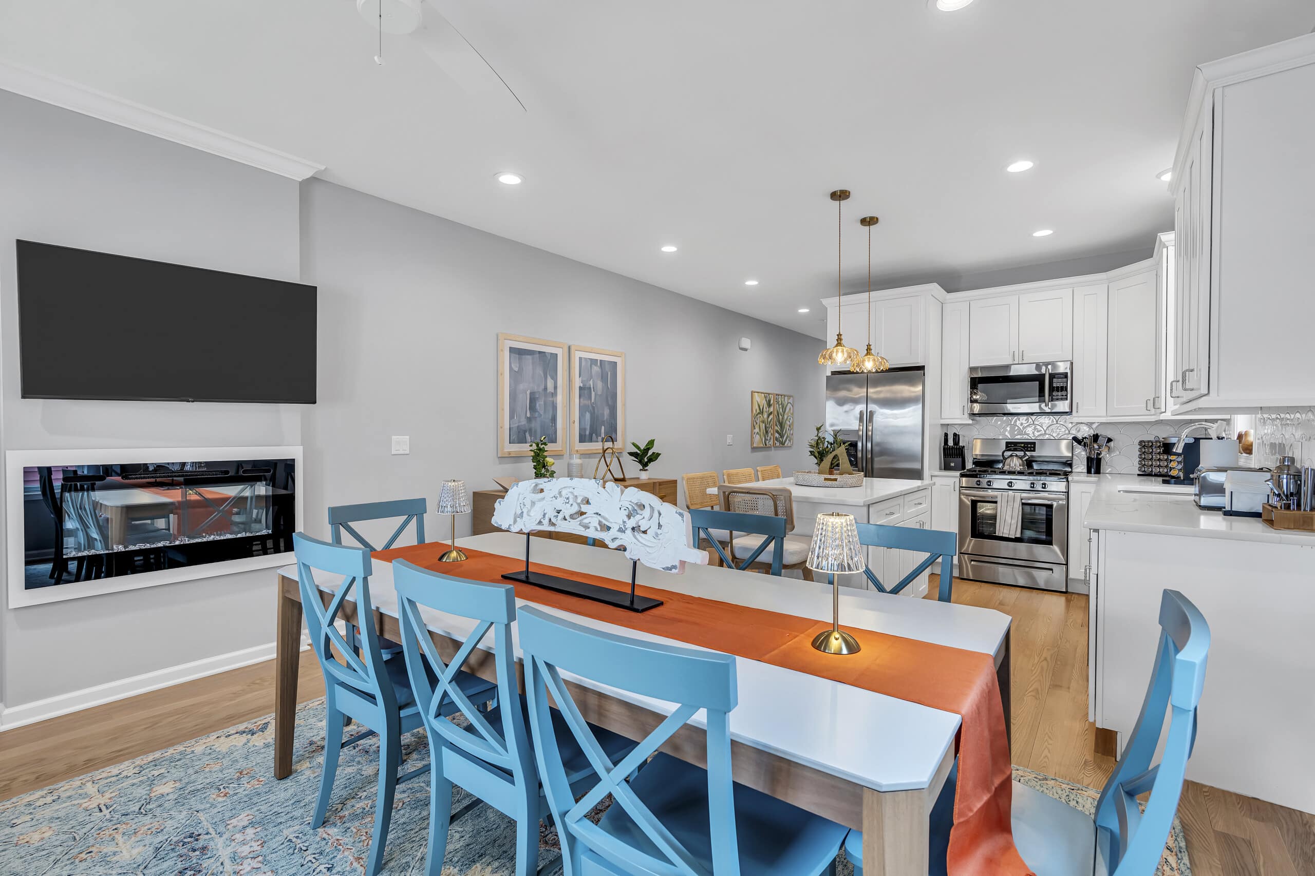 Dining area with large table, white kitchen view, two framed pictures on the wall, and a wall-mounted TV.